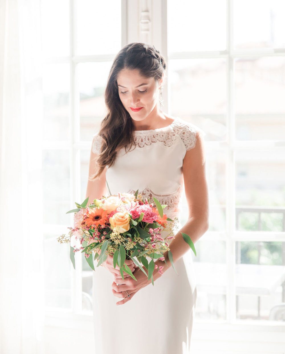 Mariée dans une robe de Rime Arodaky tenant un bouquet de fleurs devant une fenêtre