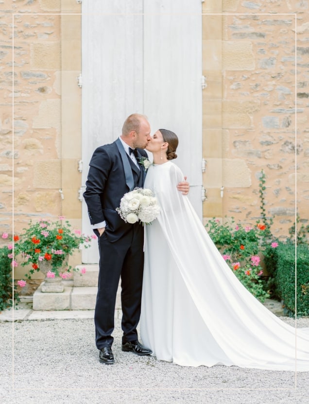 Bride and groom kissing in front of the stone wall of a french chateau in Dordogne, with some flowers behind. The groom wears a black suite and the bride a white dress with a cape