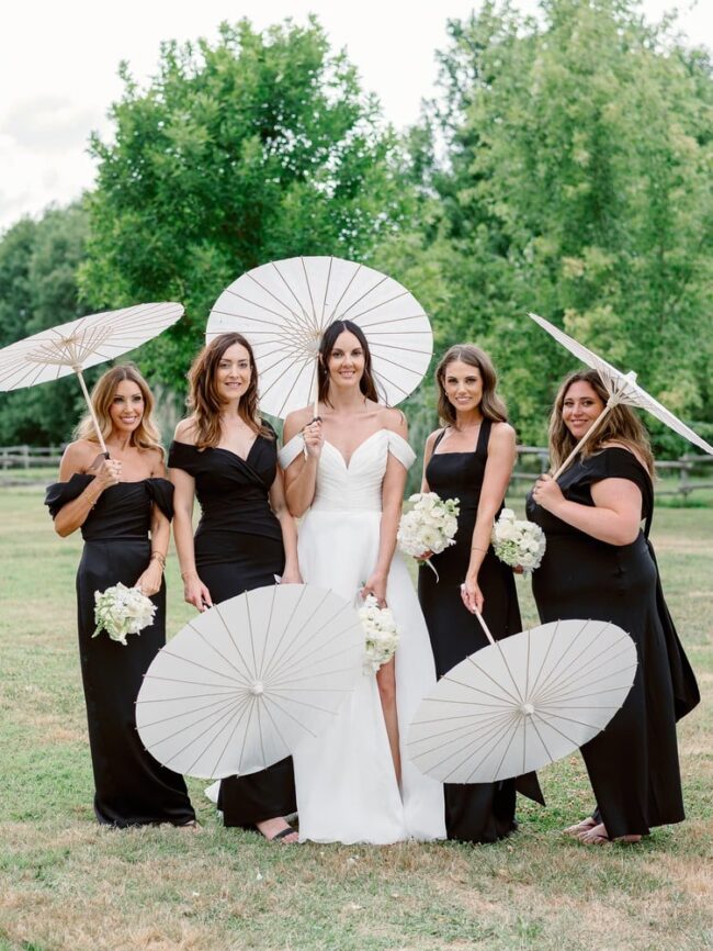 Group shot of a bride wearing a white dress and her bridesmaids wearing black dresses and holding parasols
